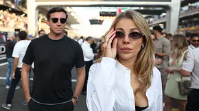 Eine stilvoll gekleidete Frau mit Sonnenbrille und weißer Bluse steht im Vordergrund auf einer belebten Rennstrecke, hinter ihr ein Mann in schwarzem T-Shirt und Sonnenbrille; im Hintergrund sind zahlreiche Zuschauer und Tribünen zu sehen. - Foto: IMAGO / NurPhoto