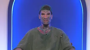 Nikola Grey sitzt mit tätowiertem Gesicht, rückwärts getragener Kappe und löchrigem Shirt vor blau beleuchtetem Hintergrund. - Foto: Joyn Nikola Grey sitzt mit tätowiertem Gesicht, rückwärts getragener Kappe und löchrigem Shirt vor blau beleuchtetem Hintergrund. - Foto: Joyn