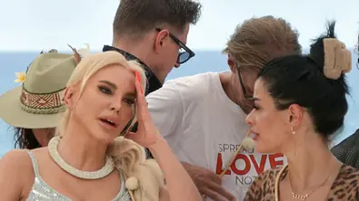 Tatjana Gsell sitzt mit blonden Haaren und glitzerndem Outfit am Set einer TV-Show, legt die Hand an die Stirn und wirkt nachdenklich. Neben ihr eine dunkelhaarige Frau im Leo-Muster-Look. Im Hintergrund sind weitere Personen, darunter ein Mann mit Brille und weißem T-Shirt, zu sehen. Die Szene spielt draußen vor hellem Himmel und Meer. - Foto: Joyn