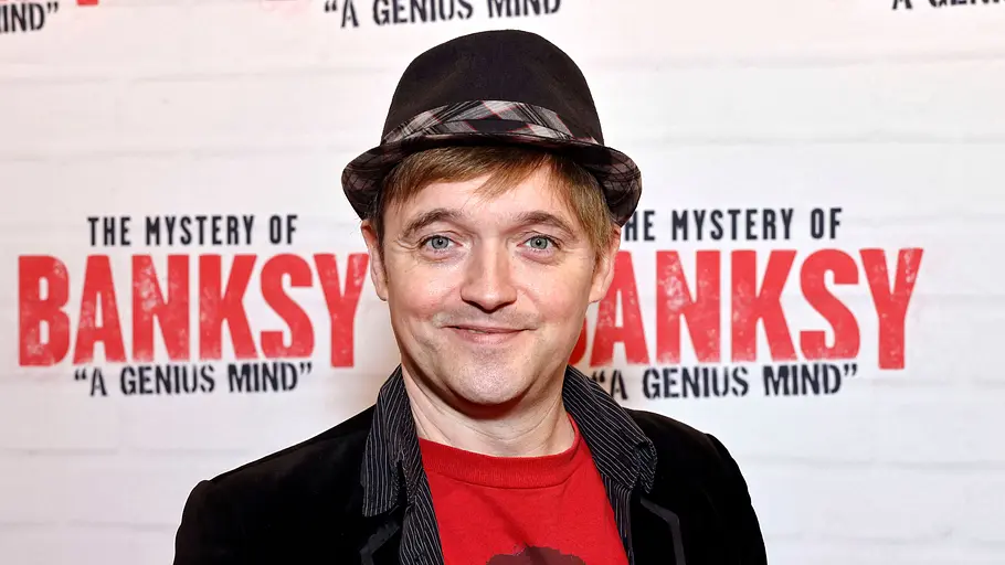 Paddy Kroetz Paddy Kroetz bei einem Event vor einer „The Mystery of Banksy“-Fotowand, trägt schwarzen Hut, rotes T‑Shirt und dunkles Sakko und lächelt in die Kamera.