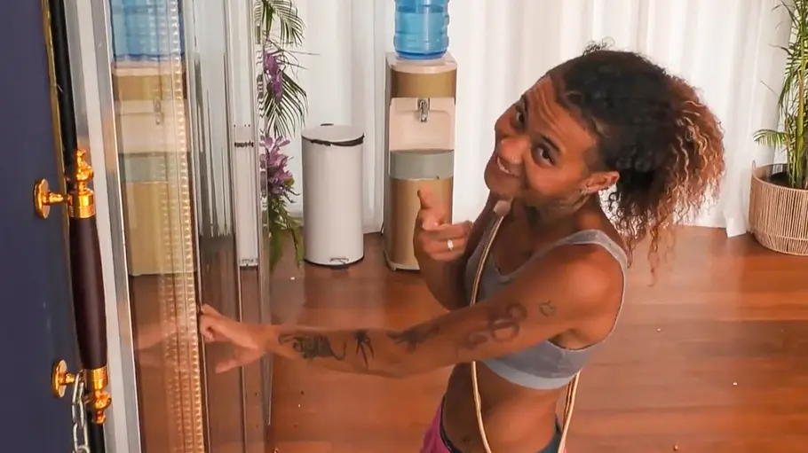 Chika Chika steht in einem Raum an einer Glastür und zeigt mit einem Finger in die Kamera; im Hintergrund sind ein Wasserspender und Pflanzen zu sehen.