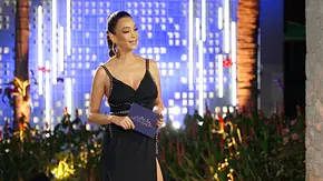 Verona Pooth steht in schwarzem Abendkleid mit hochgebundenen Haaren auf einer beleuchteten Show-Bühne und hält eine Karte in der Hand. - Foto: Joyn Verona Pooth steht in schwarzem Abendkleid mit hochgebundenen Haaren auf einer beleuchteten Show-Bühne und hält eine Karte in der Hand. - Foto: Joyn