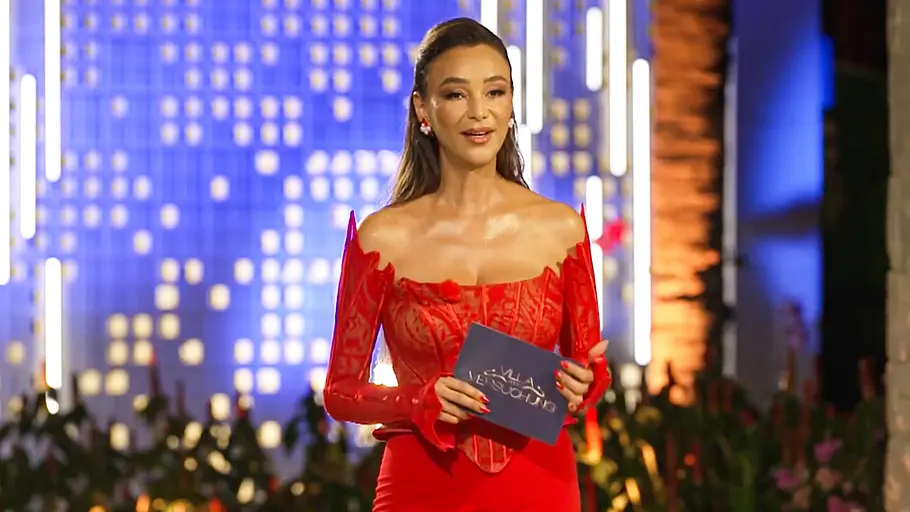 Verona Pooth Verona Pooth steht im roten Kleid auf einer beleuchteten Show-Bühne und hält eine Karte in der Hand.