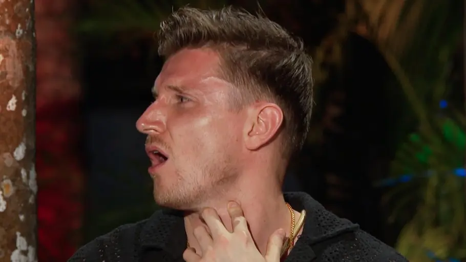 Tommy Pedroni Tommy blickt nach links und berührt seinen Hals; trägt ein schwarzes Oberteil und eine goldene Kette, im Hintergrund sind unscharfe Pflanzen und farbiges Licht zu sehen.