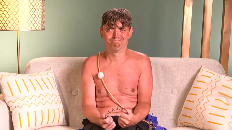 Paddy Paddy sitzt oberkörperfrei mit einem fragenden Blick und einem Haarreif in der Hand auf einem Sofa