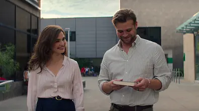 Zwei Personen stehen nebeneinander draußen auf dem Uni-Campus, die Frau lächelt den Mann an, der auf ein Buch schaut. - Foto: Courtesy of Netflix © 2026