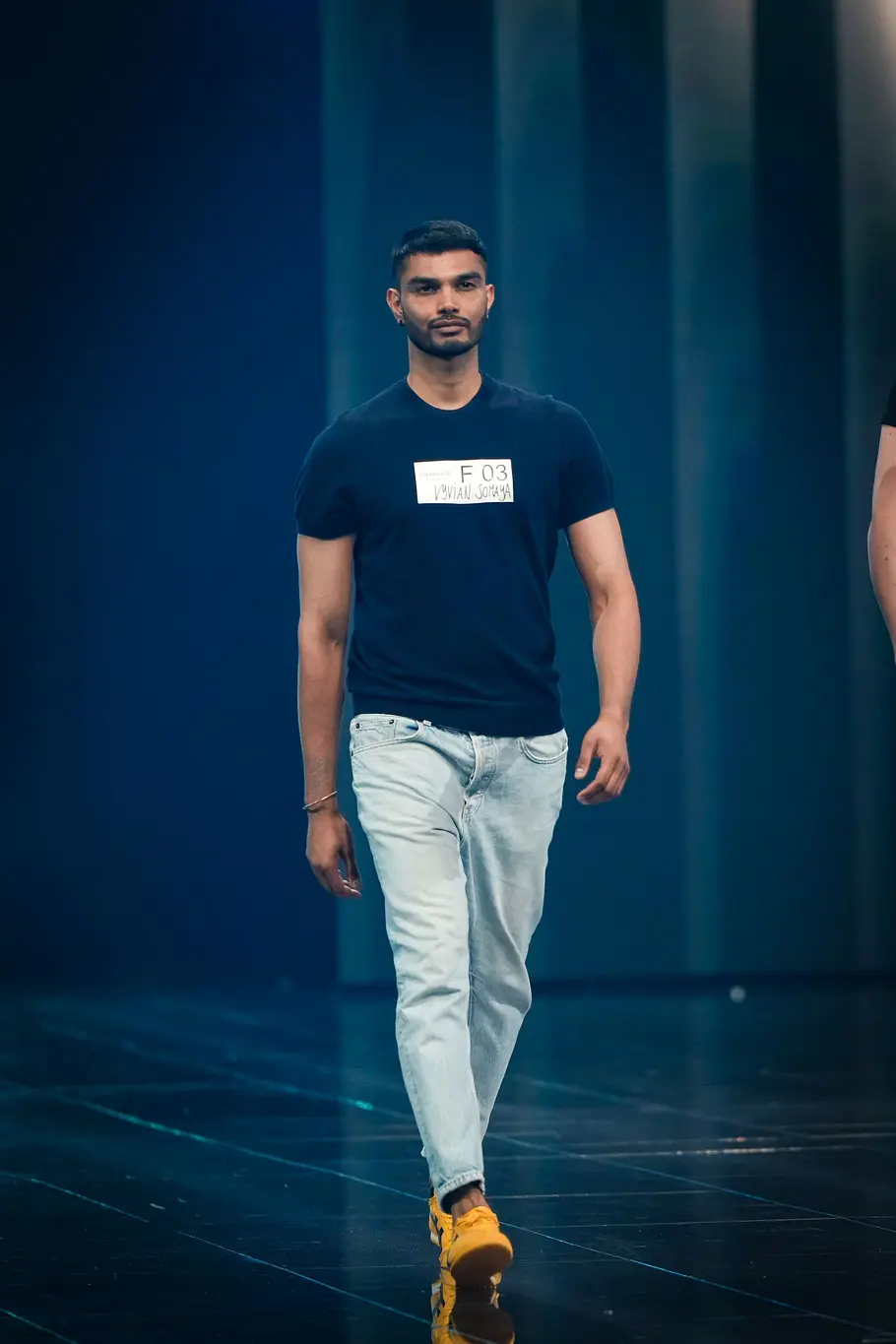 Ein Kandidat präsentiert sich beim Catwalk in schwarzem T-Shirt, heller Jeans und auffälligen gelben Schuhen vor blau beleuchtetem Hintergrund.