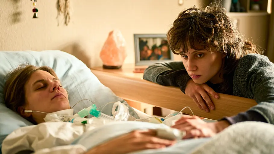Wake Up: Emilia (Luna Jordan) und Clara (Alicia von Rittberg) Clara (Alicia von Rittberg) liegt mit geschlossenen Augen in einem Krankenhausbett. An ihrem Hals befindet sich ein Schlauch, in ihren Ohren Kabelkopfhörer. Rechts neben ihr lehnt sich Emilia (Luna Jordan) über den hölzernen Bettschutz und berührt Claras linke Hand. Nachdenklich richtet Emilia ihren Blick auf die sich berührenden Hände. Im Hintergrund stehen ein Foto sowie eine Salzlampe auf einem Nachttisch.