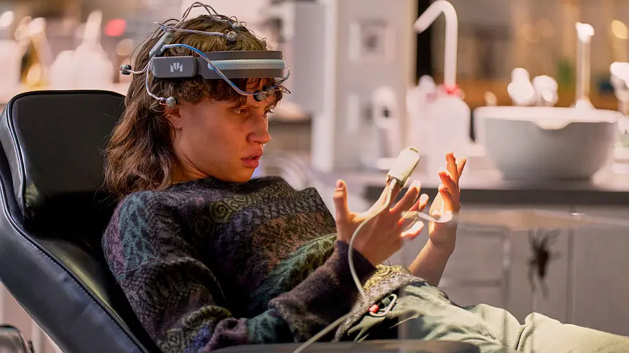 Wake Up: Emilia (Luna Jordan) Halb liegend sitzt Emilia (Luna Jordan) in einem Patientenstuhl und betrachtet ihre Hände. Im Vordergrund steht ein würfelförmiges, transparentes Terrarium mit einer großen Spinne. An Emilias rechtem Finger ist ein Pulsoxymeter befestigt. Auf ihrem Kopf trägt sie ein Haarband mit zahlreichen Sensoren.