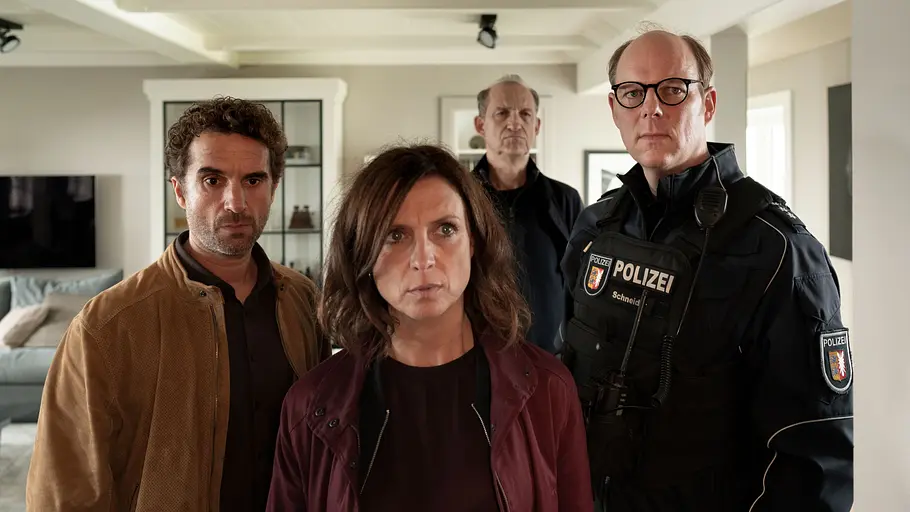 Was den Ermittlern hier wohl so große Sorgen bereitet? Unser Tipp: ein gemeingefährlicher Dackel! Hinnerk Feldmann, Ina Behrendsen, Carl Sievers und Polizist Schneider stehen in einem Wohnzimmer und schauen in die gleiche Richtung.