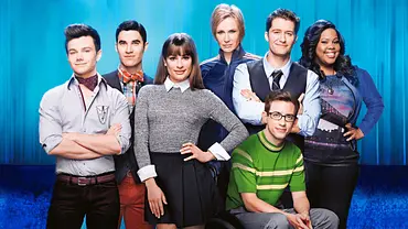 Sieben Mitglieder der „Glee“-Besetzung posieren vor blauem Hintergrund. Ein Mann mit Brille sitzt vorne im Rollstuhl. - Foto: TMDB Sieben Mitglieder der „Glee“-Besetzung posieren vor blauem Hintergrund. Ein Mann mit Brille sitzt vorne im Rollstuhl. - Foto: TMDB