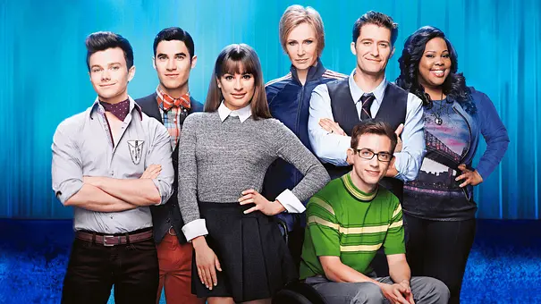 Sieben Mitglieder der „Glee“-Besetzung posieren vor blauem Hintergrund. Ein Mann mit Brille sitzt vorne im Rollstuhl. - Foto: TMDB Sieben Mitglieder der „Glee“-Besetzung posieren vor blauem Hintergrund. Ein Mann mit Brille sitzt vorne im Rollstuhl. - Foto: TMDB