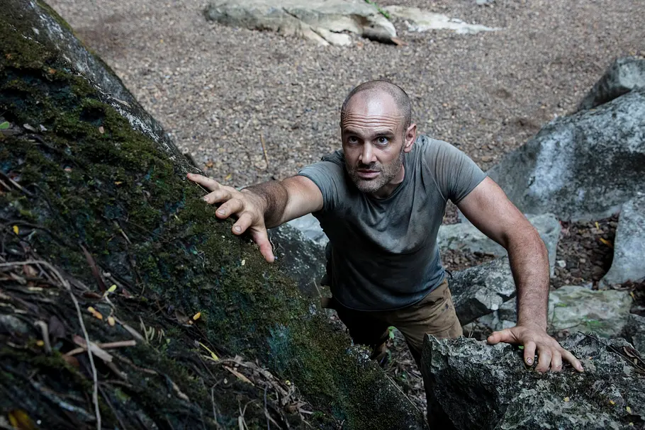 Ed Stafford klettert zwischen moosbewachsenen Felsen hinauf und stützt sich mit beiden Händen ab. Er trägt ein graues T-Shirt.