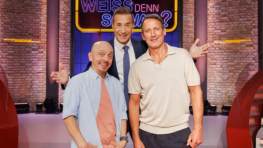 „Wer weiß denn sowas?“: Erste Gäste der neuen Staffel stehen fest – dann geht es endlich los Drei lächelnde Männer stehen im Fernsehstudio vor einem leuchtenden Schild mit der Aufschrift „Wer weiß denn sowas?“.