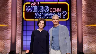 Susanne Daubner und Torsten Schröder stehen nebeneinander und lächeln vor dem leuchtenden Schriftzug „Wer weiß denn sowas?“ in einem Fernsehstudio mit Backsteinwand. - Foto: © ARD/NDR/Morric Mac Matzen