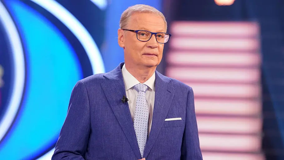 Günther Jauch Günther Jauch steht im Studio von „Wer wird Millionär?“ in einem blauen Anzug und mit Brille vor der beleuchteten Bühnenkulisse.