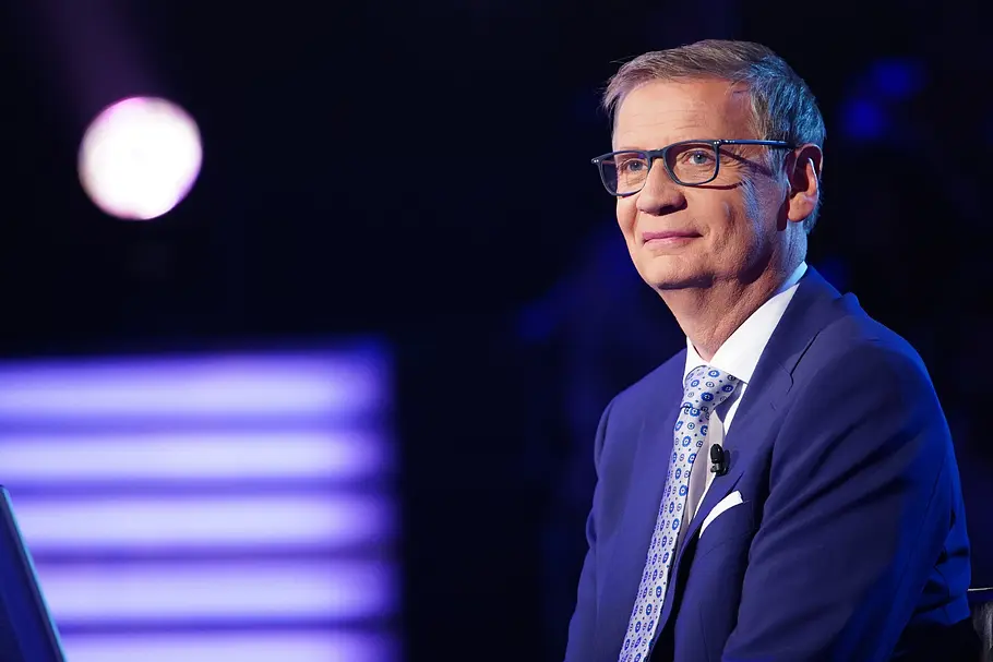 Ein Mann im blauen Anzug mit Brille sitzt in einem TV-Studio vor dunklem Hintergrund, lächelt leicht und blickt zur Seite; farbige Studioleuchten sind unscharf im Hintergrund zu sehen.