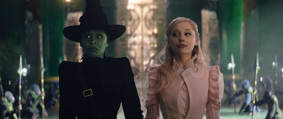 Wicked Man sieht Cynthia Erivo und Ariana Grande als Elphaba und Glinda in „Wicked“. Erivo ist grün und trägt eine schwarze Hexenrobe, während Grande ein leichtes Rosa aufträgt. Der Hintergrund ist in Smaragd-Grün. Man sieht die Affen-Wachen des Zauberers von Oz.