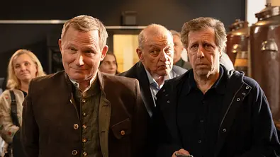 Overbeck (Roland Jankowsky) trägt Trachtenkleidung und steht neben Ekki (Oliver Korittke) im Raum einer Brauerei. Ekki hält ein Handy in der rechten Hand. Hinter den beiden steht Wilsberg (Leonard Lansink) und wirft Overbeck einen skeptischen Blick zu. - Foto: ZDF/Michael Kötschau Overbeck (Roland Jankowsky) trägt Trachtenkleidung und steht neben Ekki (Oliver Korittke) im Raum einer Brauerei. Ekki hält ein Handy in der rechten Hand. Hinter den beiden steht Wilsberg (Leonard Lansink) und wirft Overbeck einen skeptischen Blick zu. - Foto: ZDF/Michael Kötschau