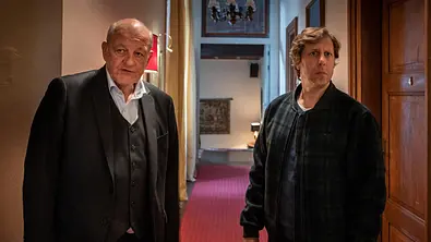 „Wilsberg – Einer von uns“: Im Flur eines Schlosses stehen Ekki (Oliver Korittke) und Wilsberg (Leonard Lansink) nebeneinander und schauen unsicher geradeaus. - Foto:  ZDF/Thomas Kost