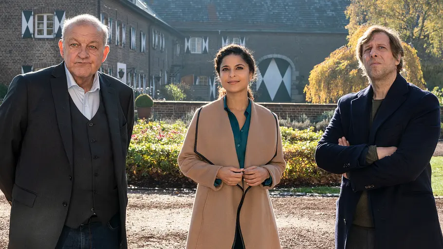 Die Anwältin Tessa Tilker (Patricia Meeden, M.) will Wilsberg (Leonard Lansink, l.) und Ekki (Oliver Korittke, r.) auf ein stilvolles Mittagessen in einem Schloss einladen. Sie glauben, sie seien die einzigen Gäste. Wilsberg - Ungebetene Gäste: Ein sonniger Herbsttag. Draußen vor einem historischen Gebäude stehen Wilsberg (Leonard Lansink), Tessa Tilker (Patricia Meeden) und Ekki (Oliver Korittke). Alle drei schauen geradeaus.