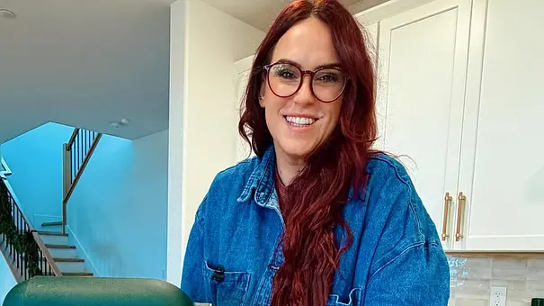 Eine Frau mit langen roten Haaren und Brille steht in einer modernen Küche und lächelt in die Kamera. - Foto: Instagram @liztomforde.author