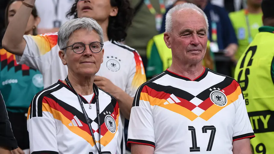 Karin Groß Zwei ältere Fans der deutschen Nationalmannschaft stehen im Stadion und tragen weiße DFB-Trikots mit der Nummer 17. Während sie konzentriert auf das Spielfeld blicken, sind im Hintergrund weitere Zuschauer, Sicherheitskräfte und Fans in Deutschland-Trikots zu sehen.