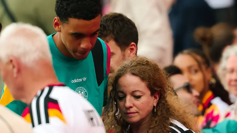 Carolin Musiala Ein Fußballspieler der deutschen Nationalmannschaft bewegt sich durch eine Menschenmenge und wird von Fans umringt. Zahlreiche Zuschauer tragen Trikots in den Farben Schwarz, Rot und Gold und hoffen auf einen kurzen Austausch oder ein Autogramm. Die Aufnahme entstand in einer belebten Stadion- oder Veranstaltungsumgebung.