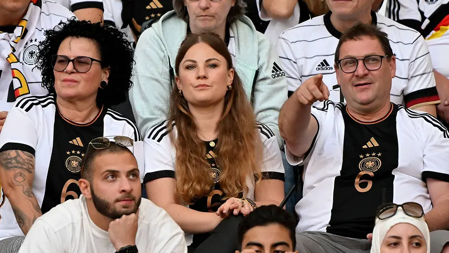 Anja Kimmich Mehrere Fußballfans sitzen auf der Tribüne eines Stadions und tragen Trikots der deutschen Nationalmannschaft. Im Mittelpunkt blickt eine junge Frau aufmerksam auf das Spielfeld, während ein Mann neben ihr gestikulierend in eine Richtung zeigt. Im Vordergrund verfolgen weitere Zuschauer das Geschehen, einige filmen oder fotografieren mit ihren Smartphones.