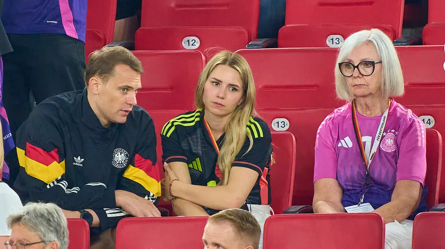 Marita Neuer Mehrere Zuschauer sitzen auf roten Tribünenplätzen in einem Fußballstadion. In der Bildmitte unterhalten sich ein Mann in einer Trainingsjacke der deutschen Nationalmannschaft und eine junge Frau im Deutschland-Trikot, während eine ältere Frau daneben aufmerksam auf das Spielfeld blickt. Im Vordergrund und Hintergrund sind weitere Fans und Besucher zu sehen.