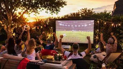 Eine Gruppe von Menschen sitzt in einem Garten vor einer großen Leinwand und schaut ein Fußballspiel, während mehrere Personen jubelnd die Arme heben. Auf einem Tisch stehen Snacks und Getränke, Lichterketten und kleine Fahnen schmücken die Umgebung. - Foto: tvmovie / mit KI erstellt Eine Gruppe von Menschen sitzt in einem Garten vor einer großen Leinwand und schaut ein Fußballspiel, während mehrere Personen jubelnd die Arme heben. Auf einem Tisch stehen Snacks und Getränke, Lichterketten und kleine Fahnen schmücken die Umgebung. - Foto: tvmovie / mit KI erstellt