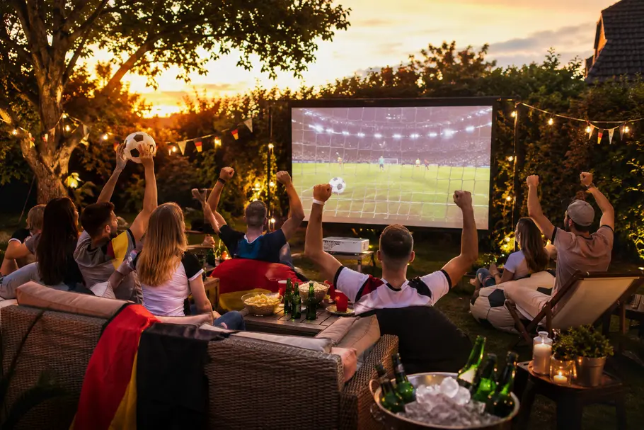 WM im Garten Eine Gruppe von Menschen sitzt in einem Garten vor einer großen Leinwand und schaut ein Fußballspiel, während mehrere Personen jubelnd die Arme heben. Auf einem Tisch stehen Snacks und Getränke, Lichterketten und kleine Fahnen schmücken die Umgebung.