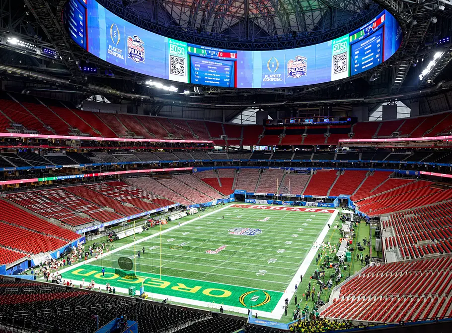 Mercedes-Benz Stadium Atlanta Innenansicht eines großen, modernen Footballstadions mit roter Bestuhlung und geschlossenem Dach; über dem Spielfeld hängt eine riesige, umlaufende Videowand mit Spielgrafiken, auf dem Feld sind Teamzonen mit „Oregon“ zu sehen und einzelne Personen stehen entlang der Seitenlinien.