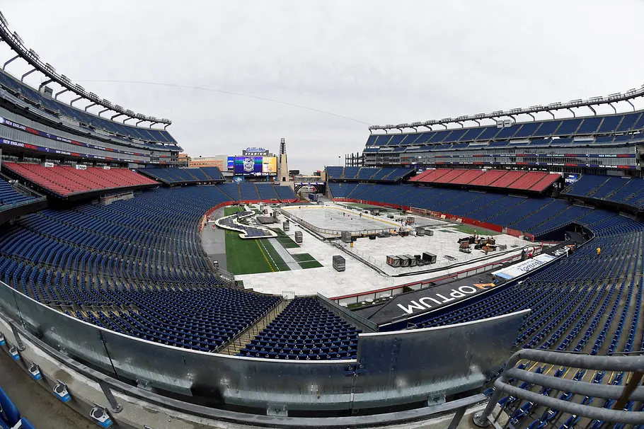 Gillette Stadium Boston Weitwinkelaufnahme eines großen, leeren Footballstadions mit blauen und roten Sitzreihen. Das Spielfeld ist größtenteils abgedeckt, in der Mitte stehen Bühnen- oder Aufbauten sowie technische Ausrüstung. Über den Rängen hängt eine große Videowand, der Himmel ist grau und bewölkt.