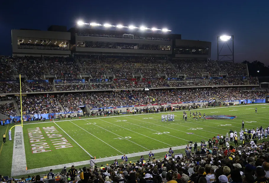 AT&T Stadium Dallas Abendaufnahme eines voll besetzten Footballstadions mit hell erleuchtetem Spielfeld; auf dem Rasen stehen zwei Teams während einer Spielunterbrechung, entlang der Seitenlinie befinden sich Trainer und Ersatzspieler, und in der Endzone ist „Pro Football Hall of Fame“ zu lesen.