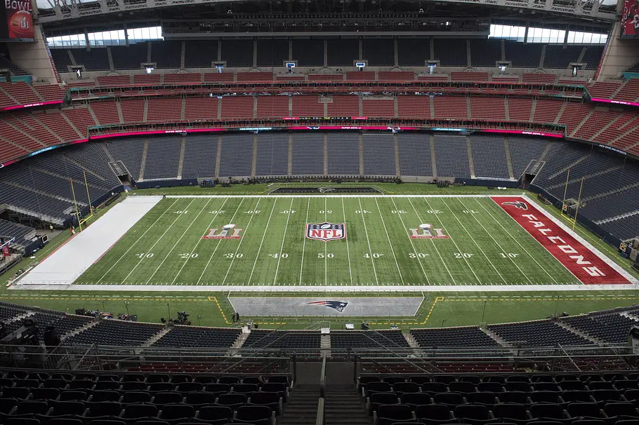NRG Stadium Houston Innenansicht eines großen, überdachten Footballstadions mit mehreren Rängen aus roten und dunklen Sitzplätzen. Auf dem Spielfeld sind das NFL-Logo sowie Super-Bowl-Markierungen zu sehen, in der Endzone steht „FALCONS“; das Stadion ist leer und für ein Spiel vorbereitet.