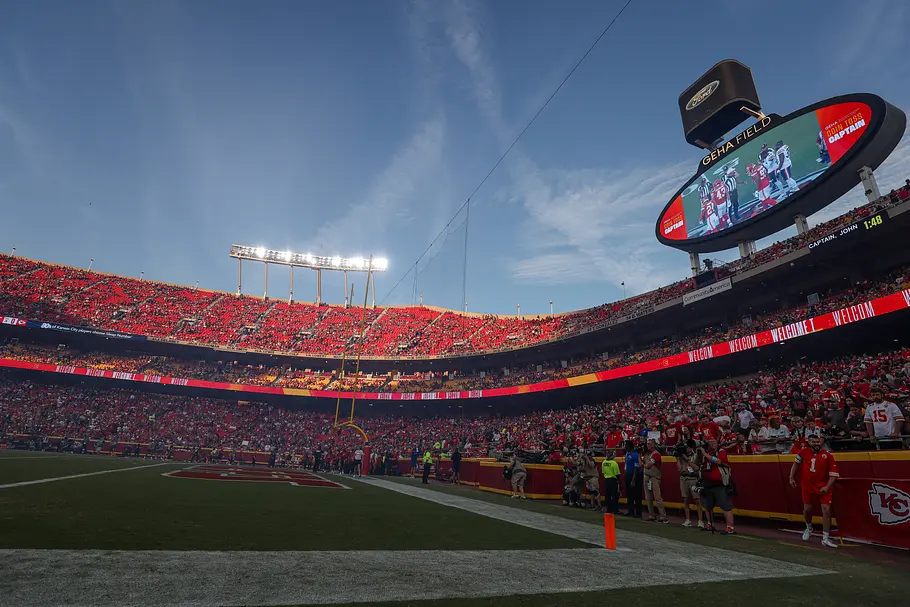 GEHA Field at Arrowhead Stadium Weitwinkelaufnahme eines voll besetzten Footballstadions während eines Spiels bei klarem Himmel. Die Ränge sind überwiegend in Rot gekleidet, auf dem Spielfeldrand stehen Fotografen und Sicherheitskräfte. Rechts ist eine große Videowand mit Spielszenen zu sehen, entlang der Tribünen leuchtet ein rotes LED-Band mit Begrüßungstexten.