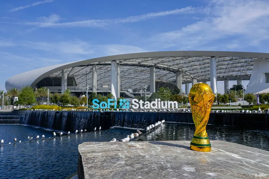 SoFi Stadium Los Angeles Goldene FIFA‑Weltmeisterschaftstrophäe im Vordergrund auf einer Steinplatte, dahinter ein Wasserbecken mit Möwen und das moderne SoFi Stadium mit geschwungenem, transparentem Dach unter blauem Himmel.