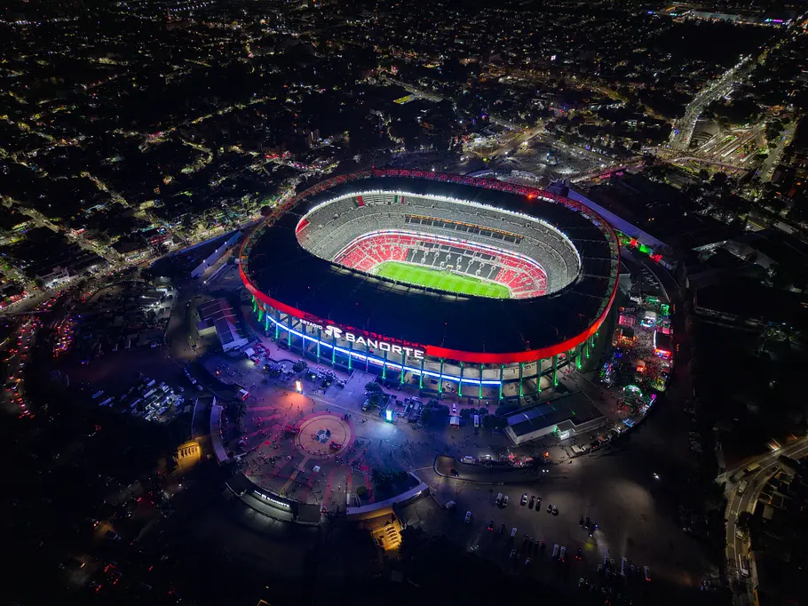 Estadio Azteca Mexico City Luftaufnahme eines beleuchteten Fußballstadions bei Nacht: Das oval geformte Stadion ist außen rot illuminiert, im Inneren strahlt das helle Spielfeld, und rundherum sind Straßen, Autos und Stadtlichter zu sehen.