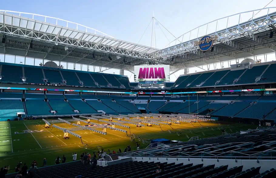 Hard Rock Stadium Miami Innenansicht des Hard Rock Stadium in Miami mit türkisfarbenen Sitzreihen und teilweise geöffnetem Dach; auf dem Spielfeld sind zahlreiche Licht- und Bühnenelemente für eine Veranstaltung aufgebaut, während auf der großen Videotafel „MIAMI“ angezeigt wird.