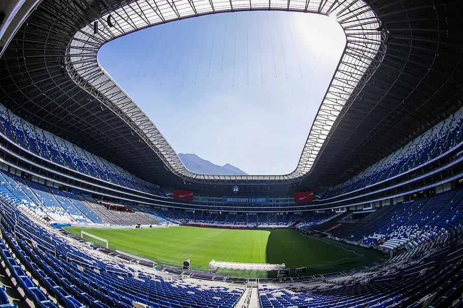 Estadio BBVA Monterrey Weitwinkelaufnahme eines modernen Fußballstadions mit blau‑weißen Sitzreihen und großem, ovalem Dach mit offener Mitte; das grüne Spielfeld liegt im Schatten, und durch die Dachöffnung sind Berge im Hintergrund sichtbar.