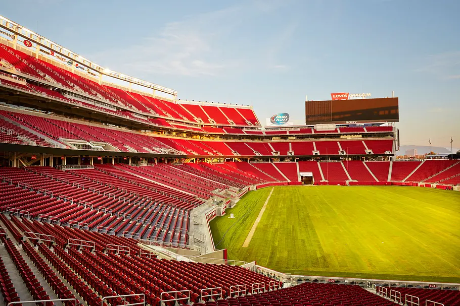 Levis Stadium San Francisco Innenansicht eines großen Footballstadions mit leeren roten Sitzreihen, die sich in mehreren Ebenen um das grüne Spielfeld ziehen; gegenüber befindet sich eine große Videotafel mit der Aufschrift „Levi’s Stadium“, während die Abendsonne die Tribünen beleuchtet.