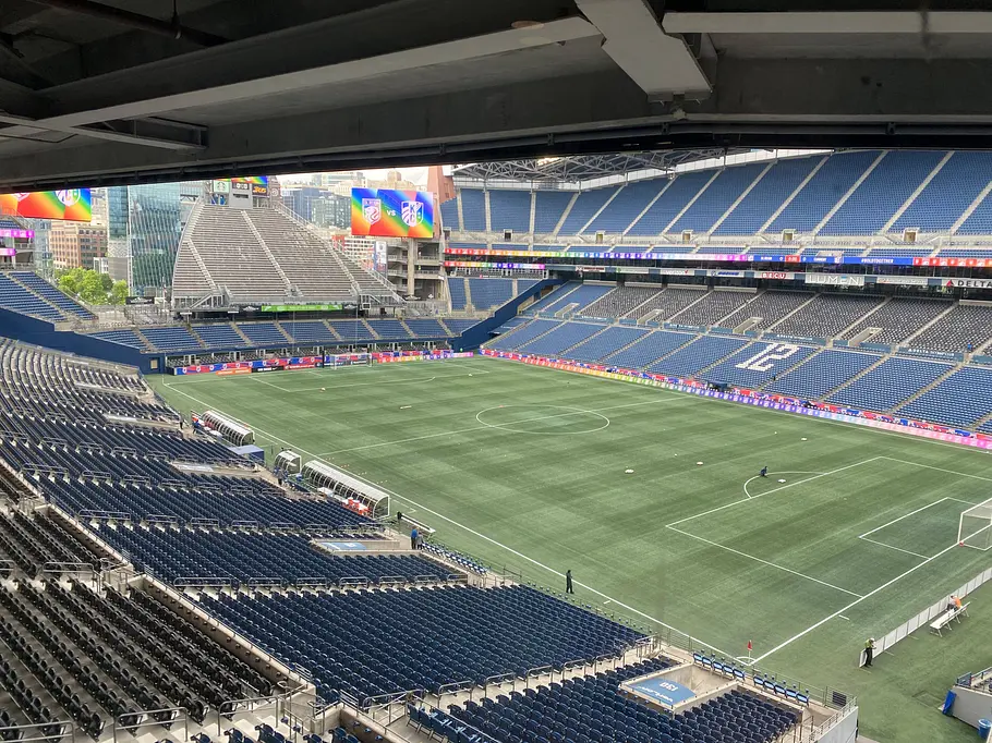 Lumen Field Seattle Innenansicht eines großen, modernen Stadions mit überwiegend blauen Sitzreihen und teilweise überdachter Tribüne; auf dem grünen Spielfeld sind Fußballmarkierungen zu sehen, während Anzeigetafeln und Werbebanner entlang der Ränge angebracht sind, die Zuschauerränge sind weitgehend leer.