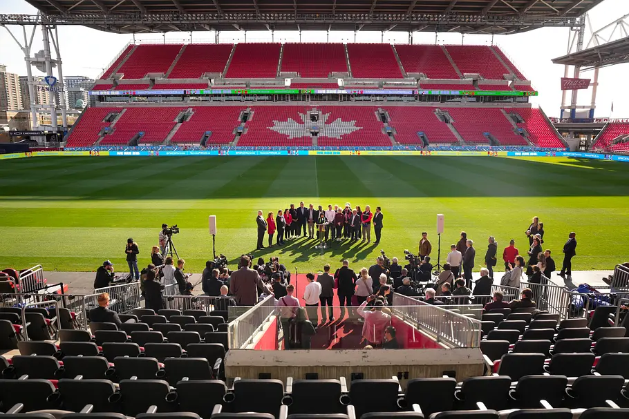 BMO Field Toronto Presseveranstaltung auf einem Fußballfeld: Eine Gruppe von Personen steht im Mittelpunkt des Rasens, umgeben von Kamerateams und Fotografen, während im Hintergrund rote Tribünen mit einem großen weißen Ahornblatt zu sehen sind.