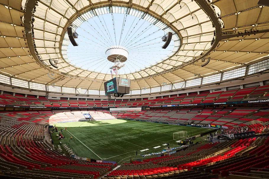 BC Place Stadium Vancouver Innenansicht eines modernen Fußballstadions mit roten und grauen Sitzreihen, teilweise überdachtem Rang und offenem Dachring in der Mitte. Über dem Spielfeld hängt eine zentrale Videowürfel-Anzeige, während das grüne, leere Spielfeld von Werbebanden umgeben ist; nur wenige Personen befinden sich am Spielfeldrand.