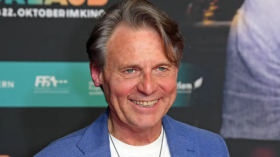 Wolfgang Bahro Lets Dance Wolfgang Bahro lächelt vor einer Filmplakatwand. Er trägt ein blaues Sakko über einem weißen Shirt und eine schwarze Kette.
