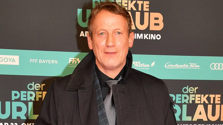 Wotan Wilke Möhring steht in schwarzem Mantel und grauer Krawatte vor einer Werbewand für „Der perfekte Urlaub“.