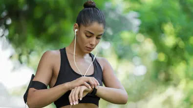 Eine sportliche Frau mit Kabelkopfhörern schaut während einer Sporteinheit auf die Smartwatch an ihrem Handgelenk. - Foto: TVMovie/Canva