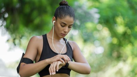 Eine sportliche Frau mit Kabelkopfhörern schaut während einer Sporteinheit auf die Smartwatch an ihrem Handgelenk. - Foto: TVMovie/Canva