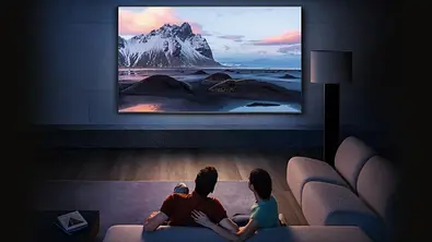 Ein großer Fernseher mit Schneelandschaft hängt in einem Wohnzimmer. Davor ein Paar auf dem Sofa. - Foto: amazon/PR
